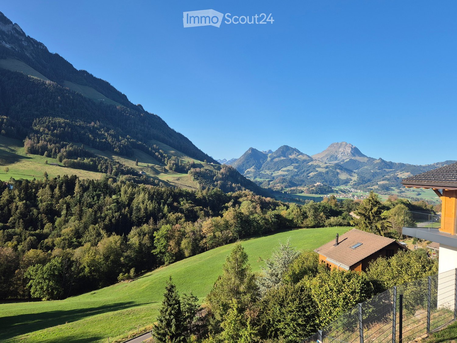 A scenic view of a lush green valley surrounded by dense forests and towering mountains under a clear blue sky. A small house with a red-tiled roof is visible in the foreground, nestled among trees.