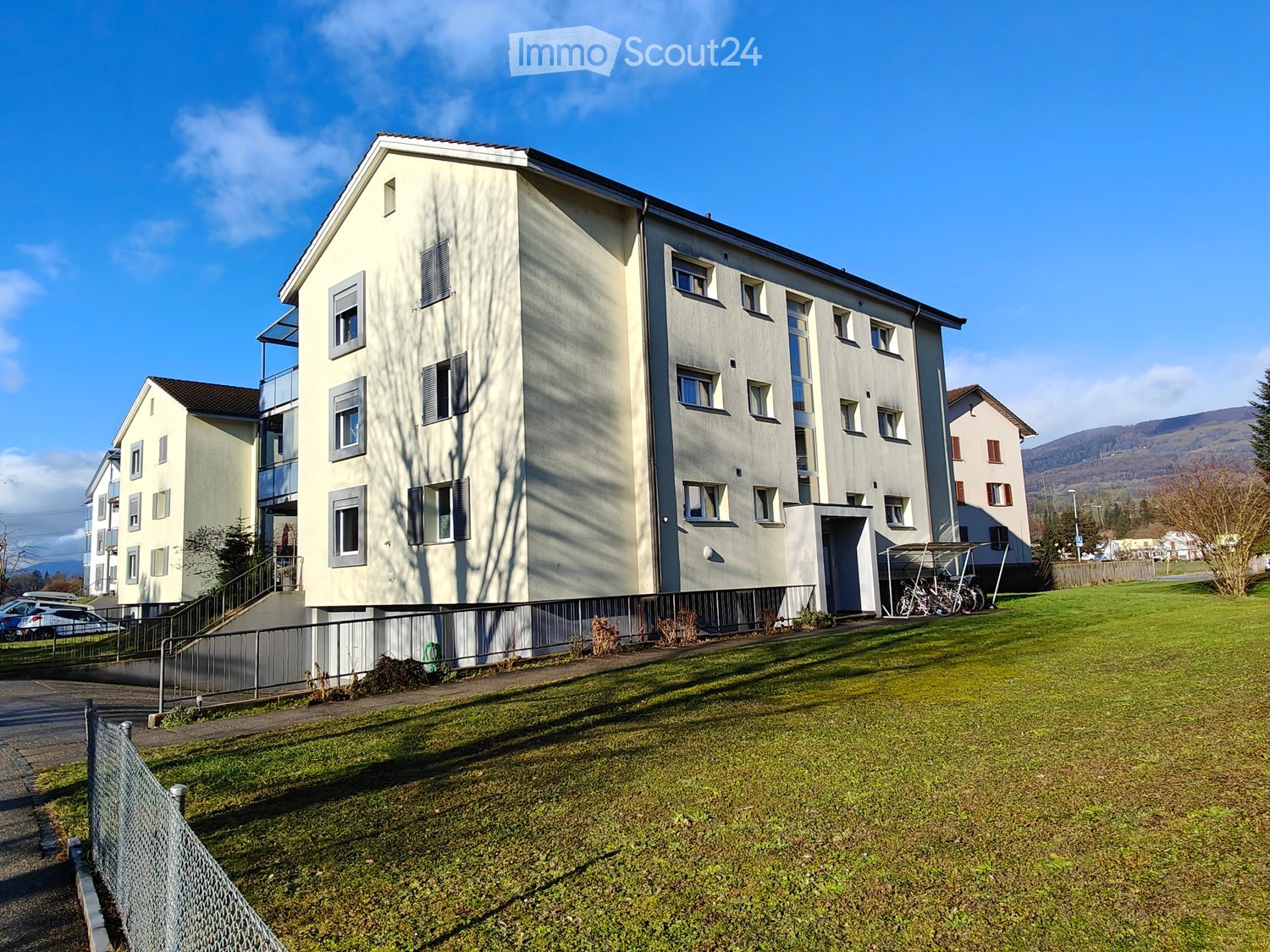 Multi-story residential building with light-colored facade, multiple windows, and balconies. The building is surrounded by a grassy area with a fence and a few parked bicycles.