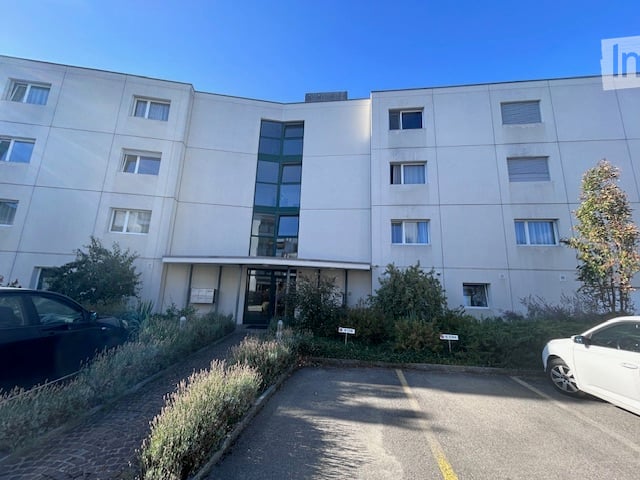 Modern multi-story apartment building with white facade, large windows, and a central entrance. There is a paved parking area in front with two cars parked.