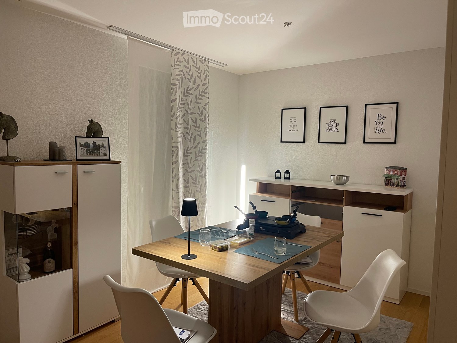 A modern, well-lit room with a wooden table, white chairs, a cabinet, and a small induction stove. The room features framed artwork, a lamp, and a small cabinet with decorative items.