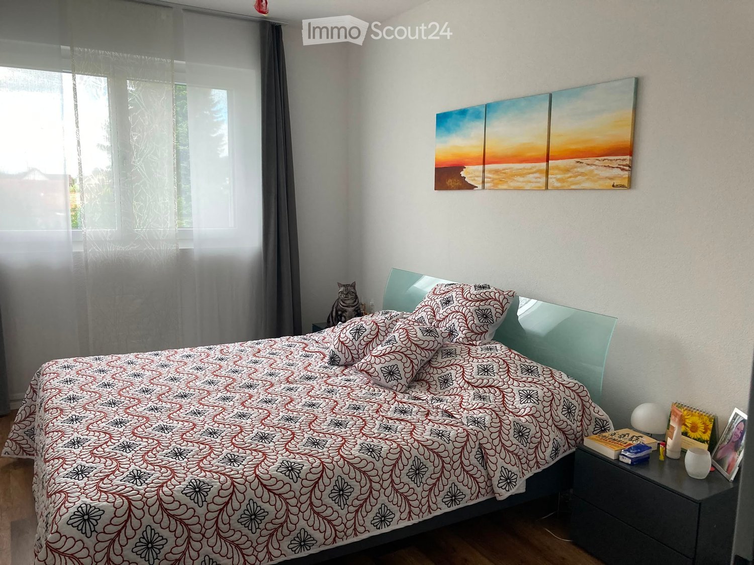A well-lit bedroom with a large bed, patterned bedding, a nightstand with a lamp, a cat, and a scenic painting on the wall.