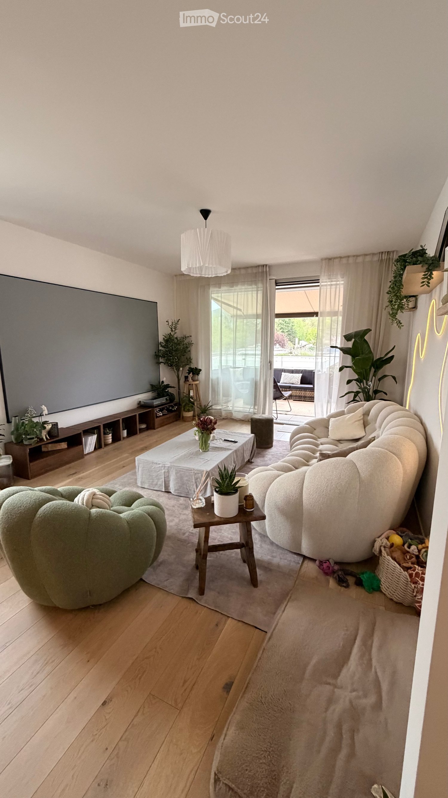 Modern living room with wooden flooring, large window, white sofa, coffee table, ottoman, side table, potted plants, and a TV screen.