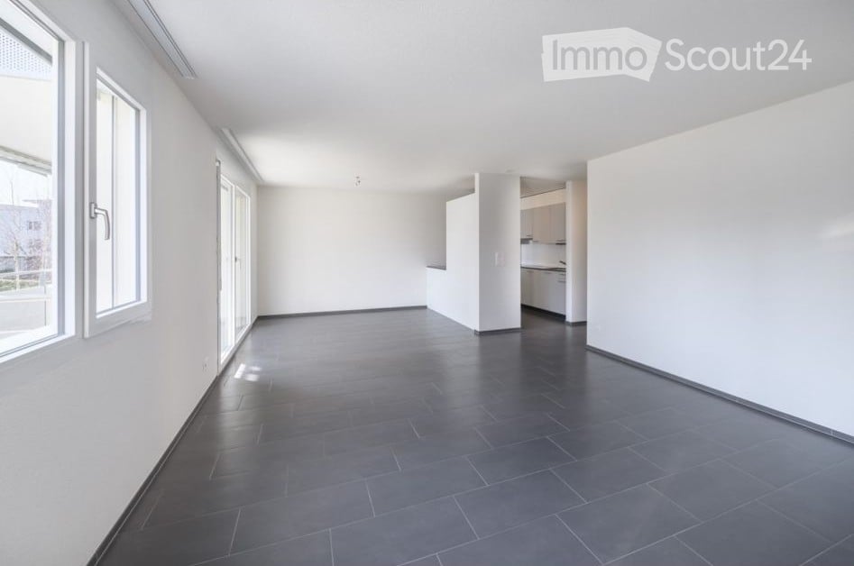 Empty room with white walls, dark tiled floor, large windows, and a small kitchen area with cabinets and appliances visible in the background.