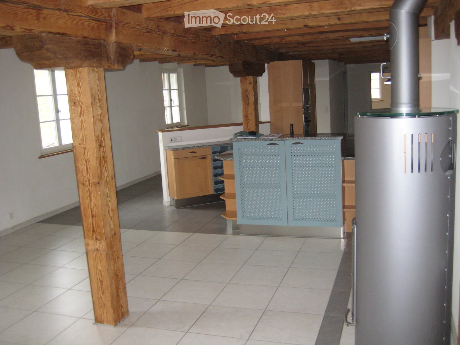 Open-plan space with exposed wooden beams, tiled floor, light-colored cabinets, and a countertop with a stool.