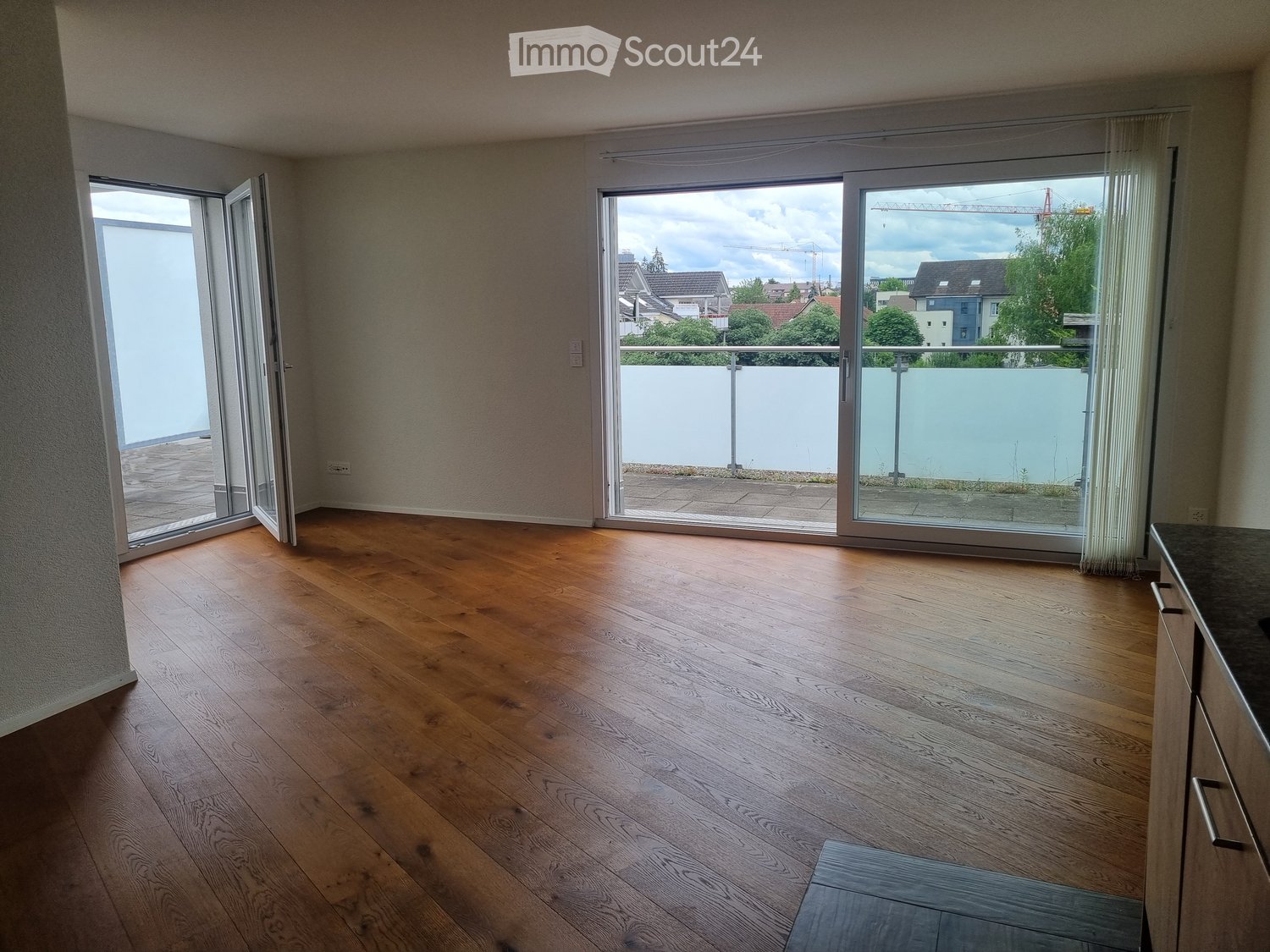 Empty room with wooden flooring, large sliding glass doors leading to a balcony, and a view of the surrounding area.