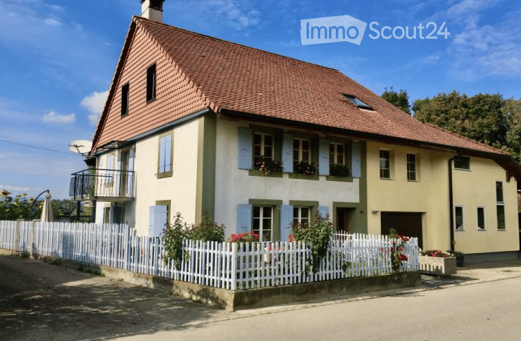 Two-story house with a red-tiled roof, light yellow walls, and blue shutters. Features a small balcony on the upper floor, a white picket fence, and a garden with flowers.