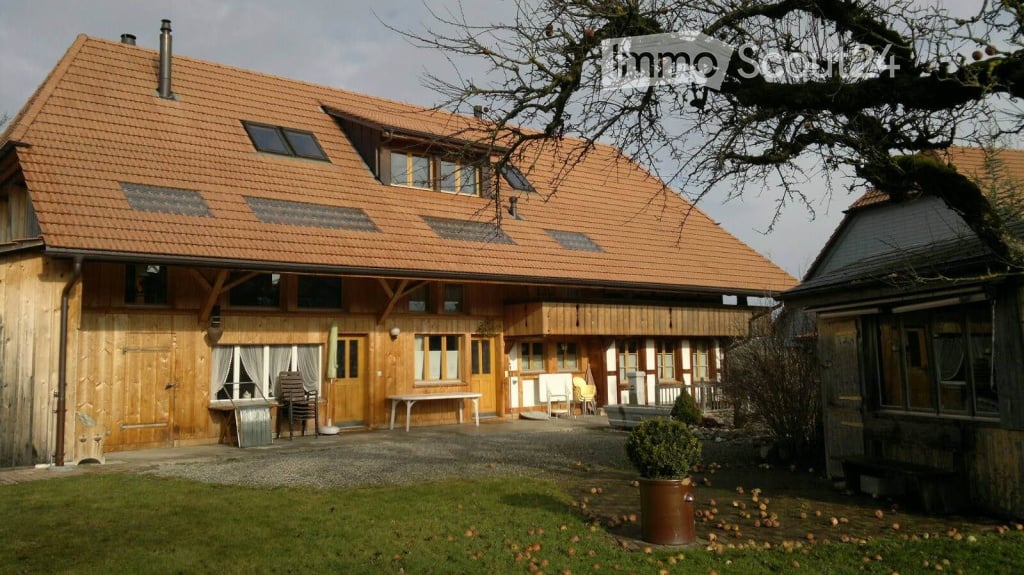 Two-story wooden house with a red-tiled roof, multiple windows, and a covered porch area with outdoor furniture.