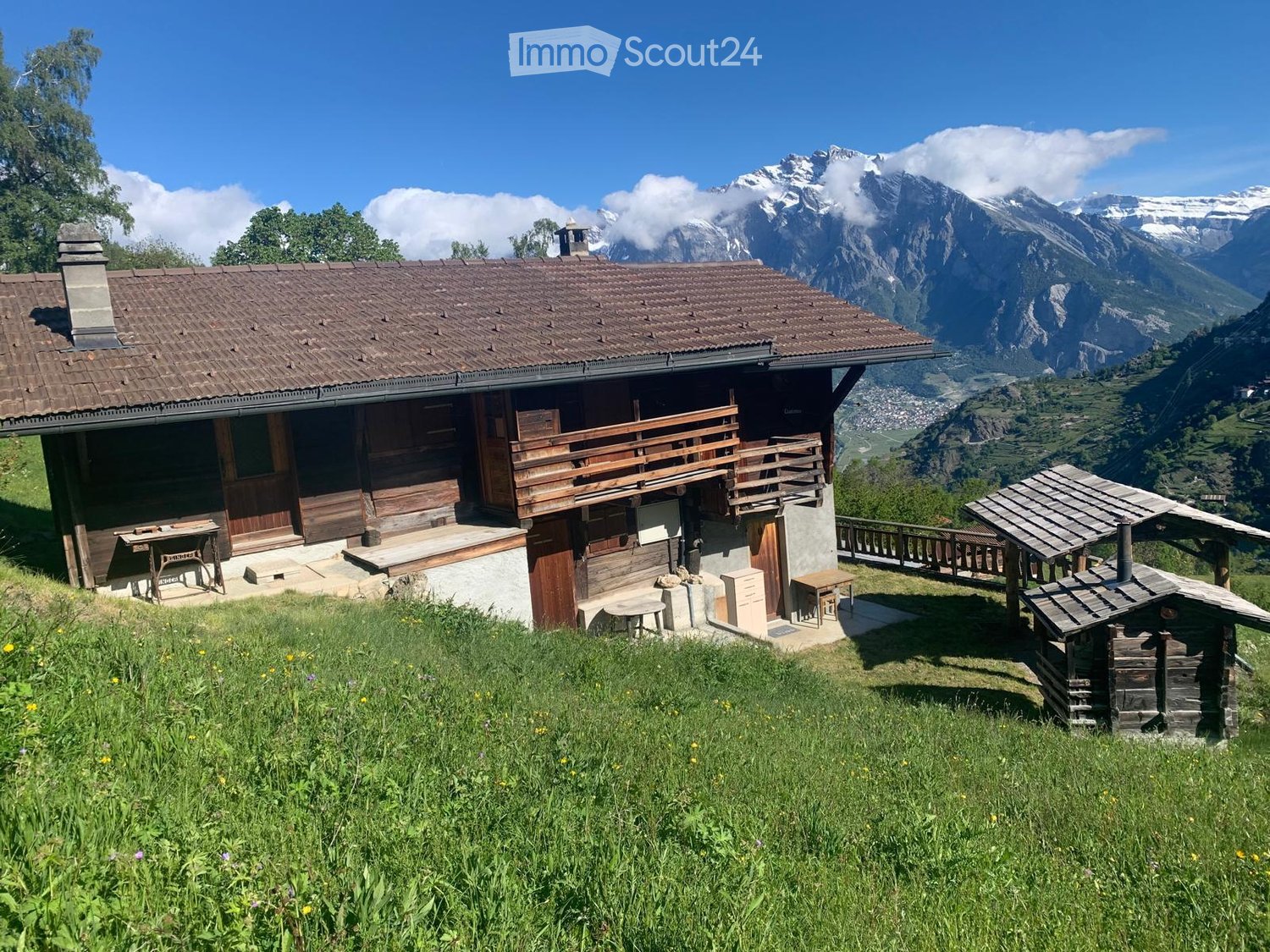 A wooden chalet with a brown tiled roof, wooden balcony, and stone base, situated on a grassy slope with a stunning mountain backdrop.