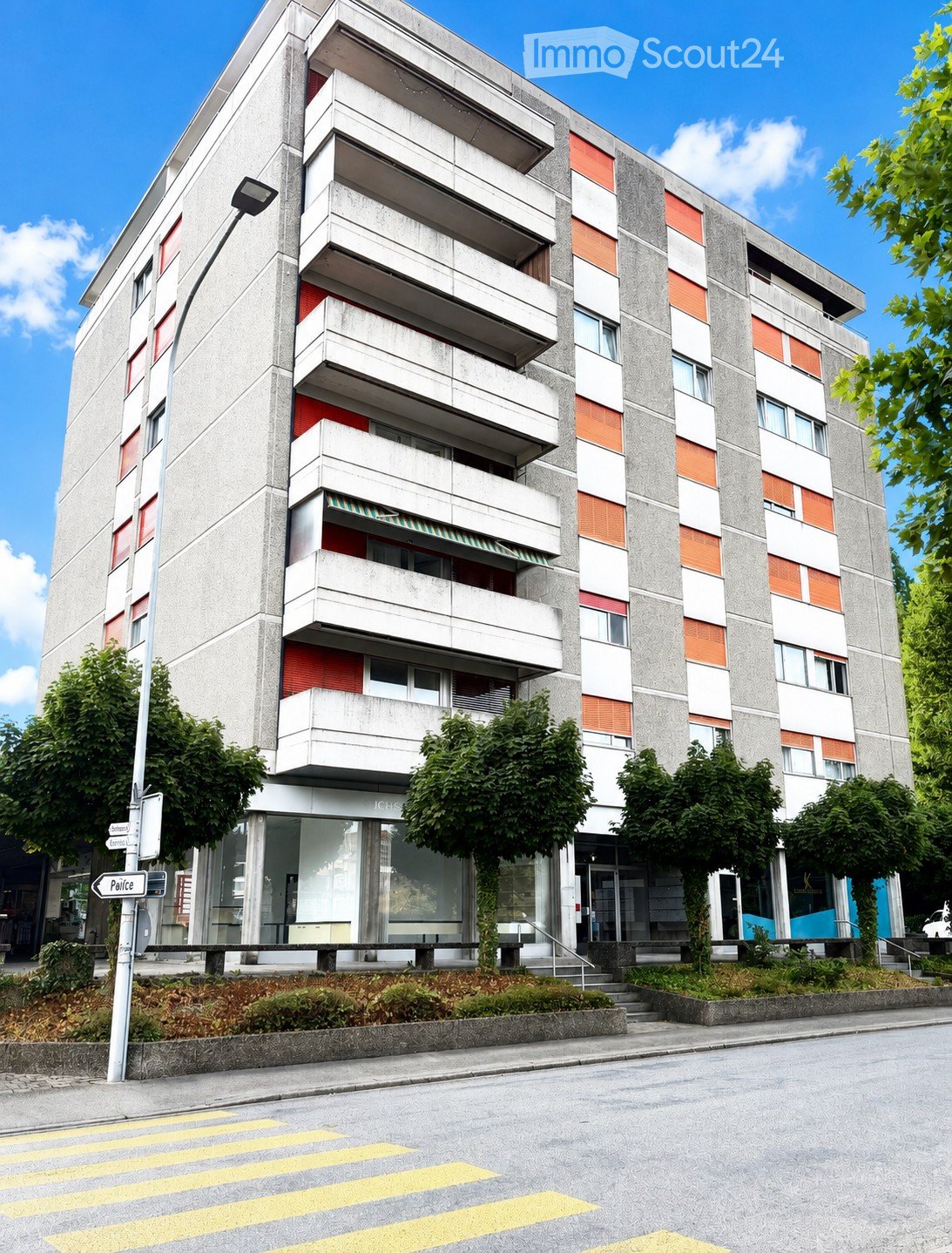 Modern multi-story apartment building with concrete and red brick facade, balconies, and greenery around the base.