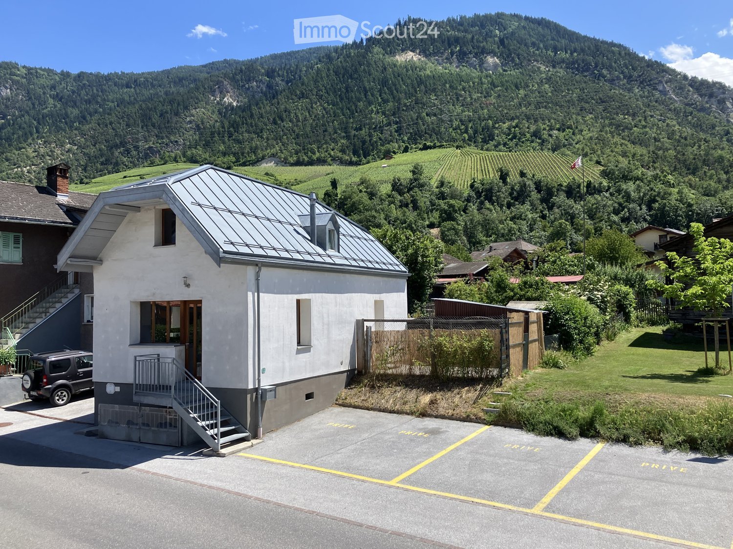 Single-story house with a grey metal roof, white walls, and a small staircase leading to the entrance. The house is situated in a scenic valley with vineyards and mountains in the background.