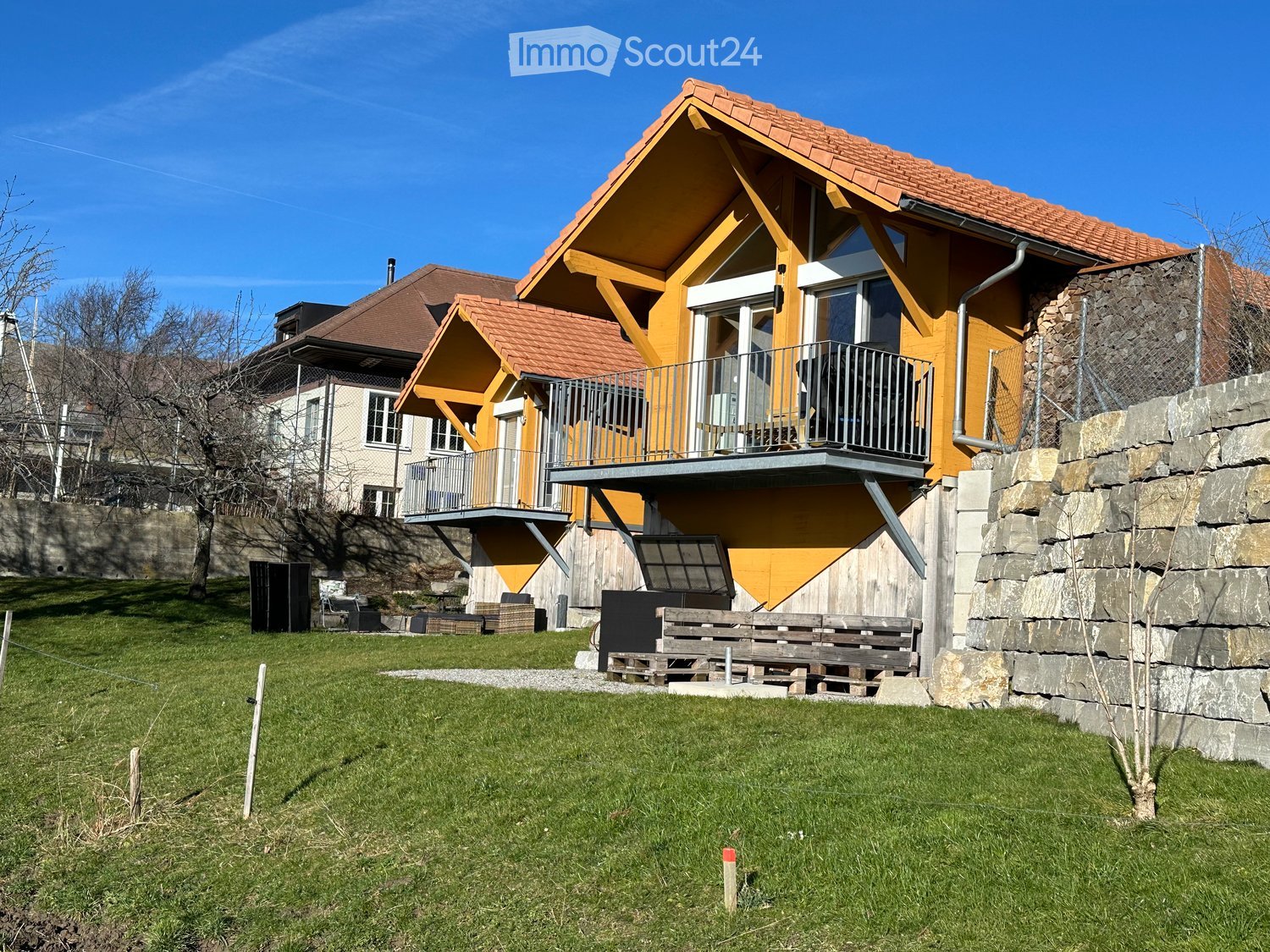 Modern two-story house with yellow exterior, red-tiled roof, and balcony overlooking a grassy yard.