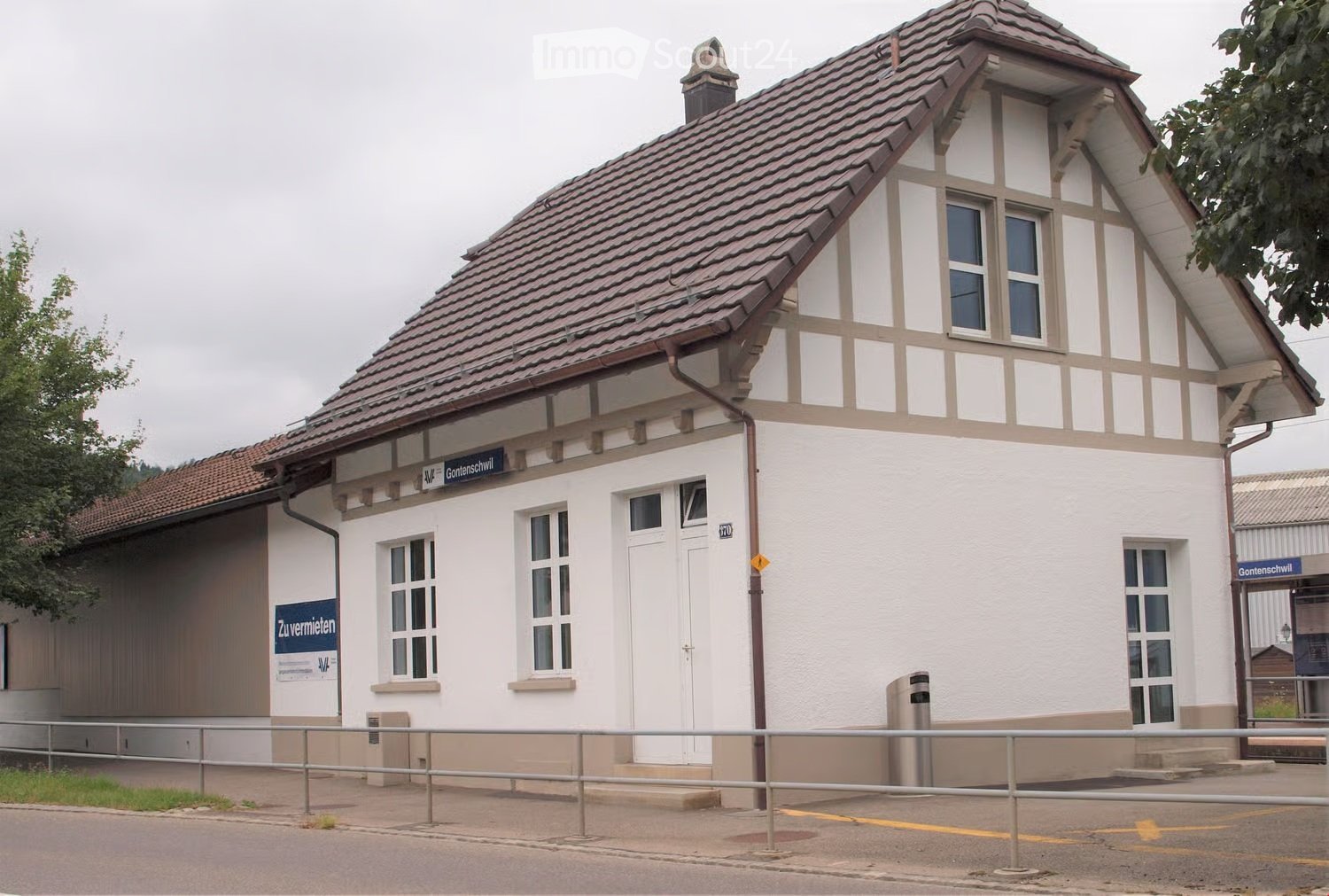 Two-story building with a tiled roof, white walls, and multiple windows. Features a sign reading "Gontenschwil" and a small entrance area with steps.