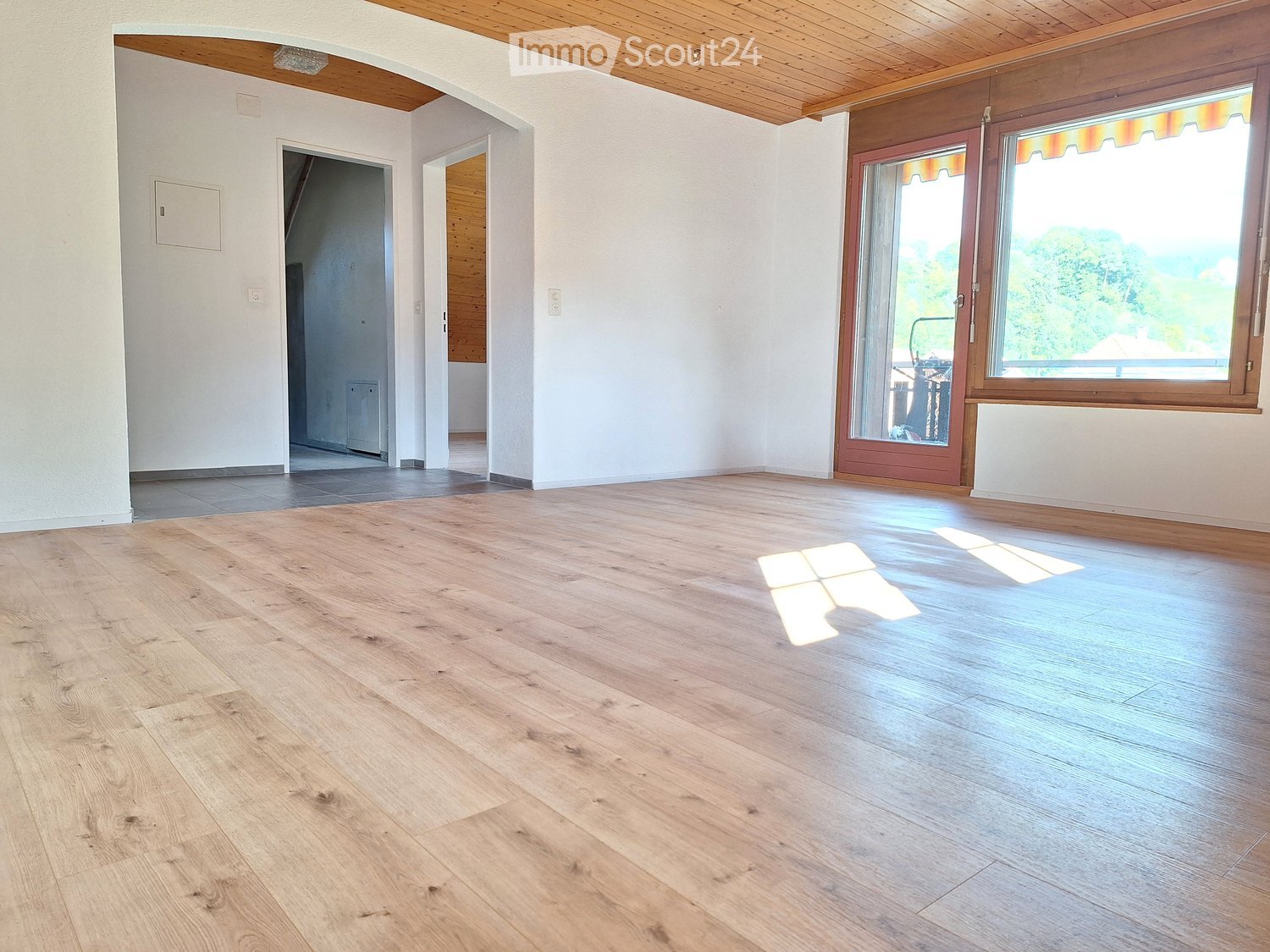 Empty room with wooden flooring, white walls, and two doors leading to other rooms. There is a balcony with a view of trees and a house outside.