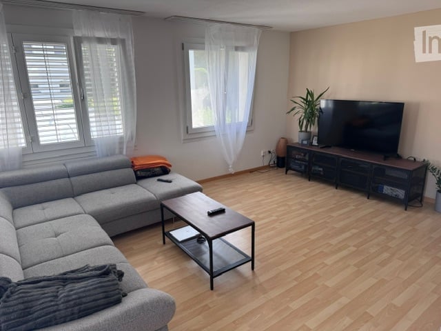 Modern living room with grey sectional sofa, coffee table, TV stand, and a potted plant. The room features large windows with white blinds and wooden flooring.