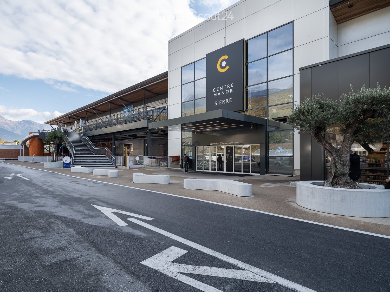 Modern building entrance with glass facade, sign reading "CENTRE MANOR SIERRE", steps leading up, and a tree in a planter.
