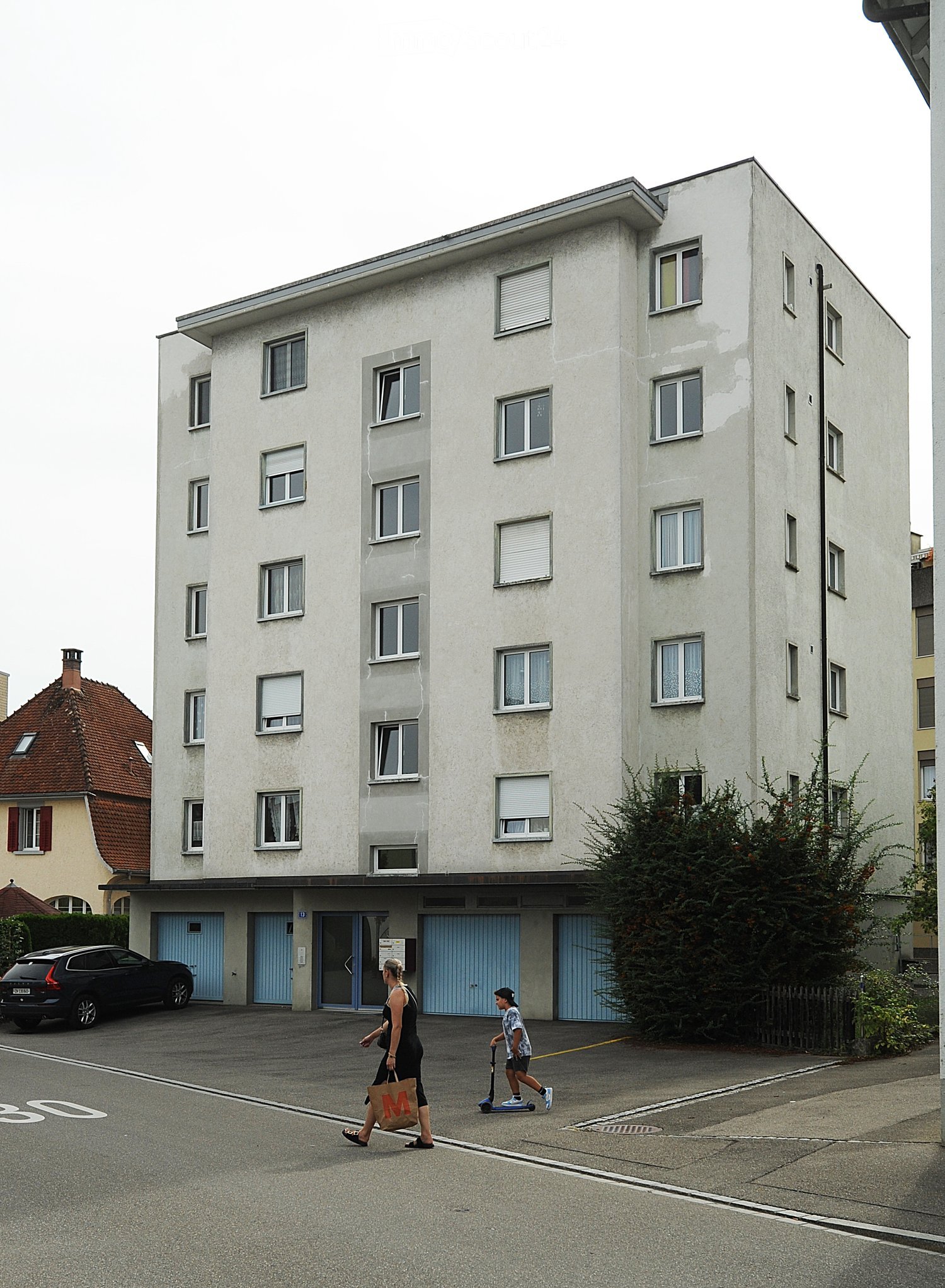 A multi-story residential building with light-colored walls and multiple windows, featuring a parking area in front with a car and two people walking.