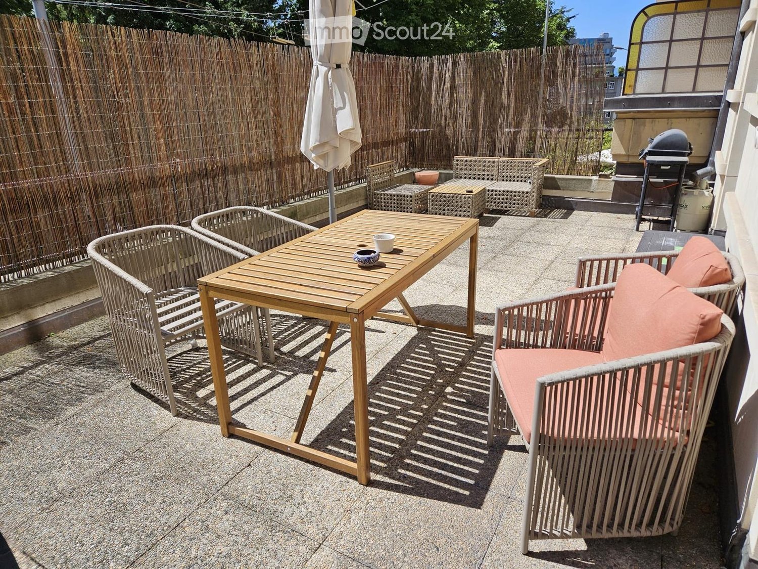 A small outdoor patio with a wooden table, wicker chairs, and an umbrella, enclosed by a bamboo fence.
