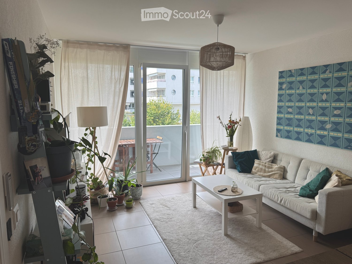 Bright living room with large sliding glass doors leading to a balcony, white sofa with cushions, coffee table, floor lamp, and several potted plants.