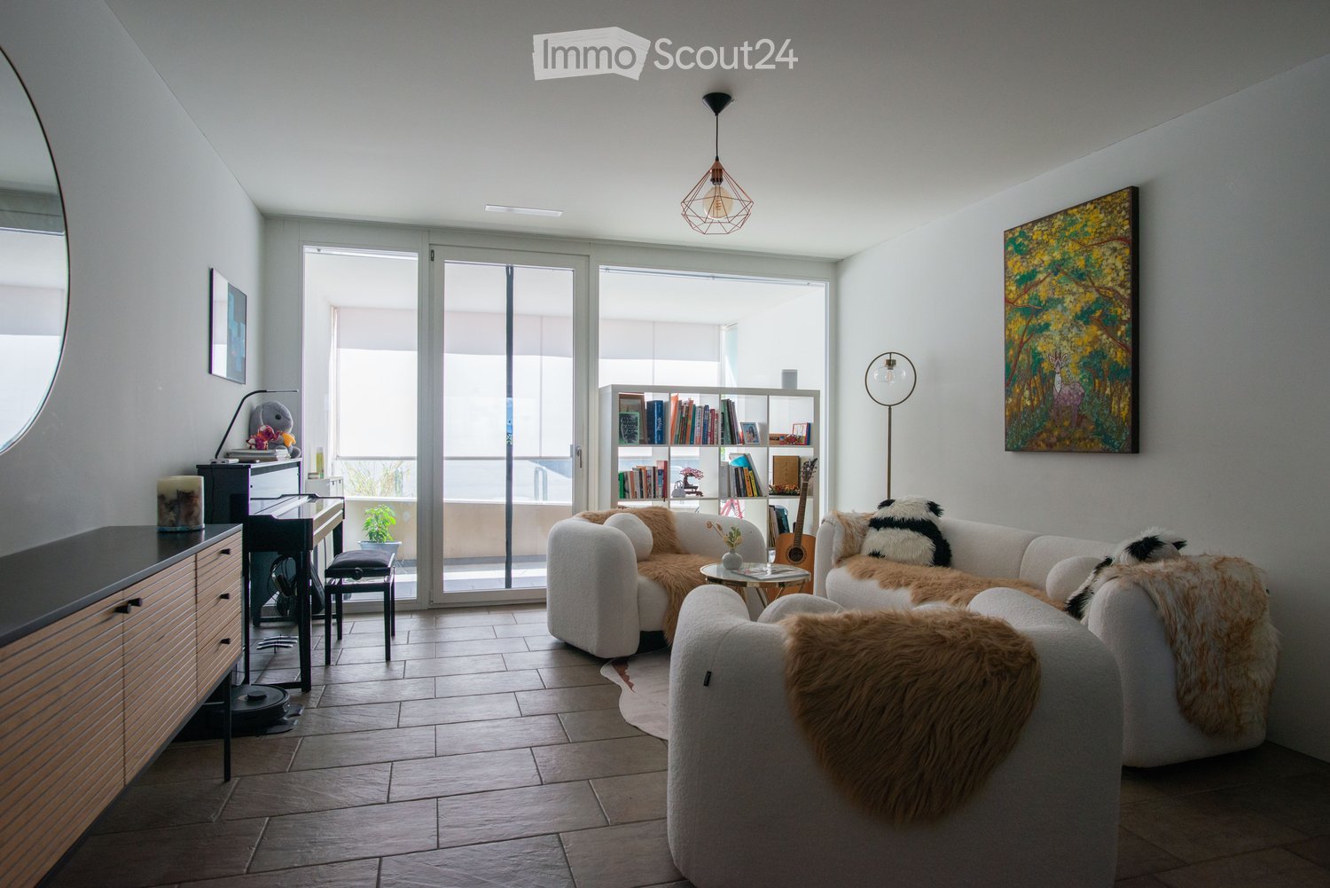 Modern living room with large sliding glass doors leading to a balcony, white sectional sofa with fur throws, coffee table, bookshelf, guitar, and a painting on the wall.