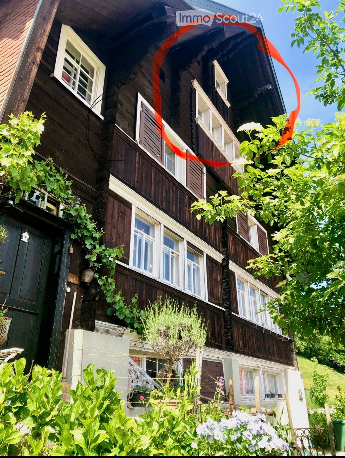 A two-story wooden house with white-framed windows, wooden shutters, and a balcony on the upper floor. The house is surrounded by lush greenery and flowering plants.
