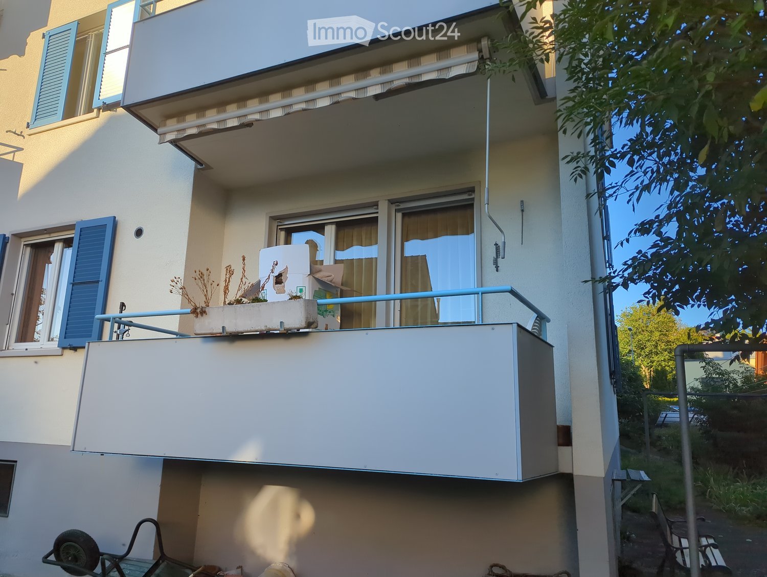 Balcony with a flower pot and a railing, attached to a multi-story building with blue shutters and a covered section.