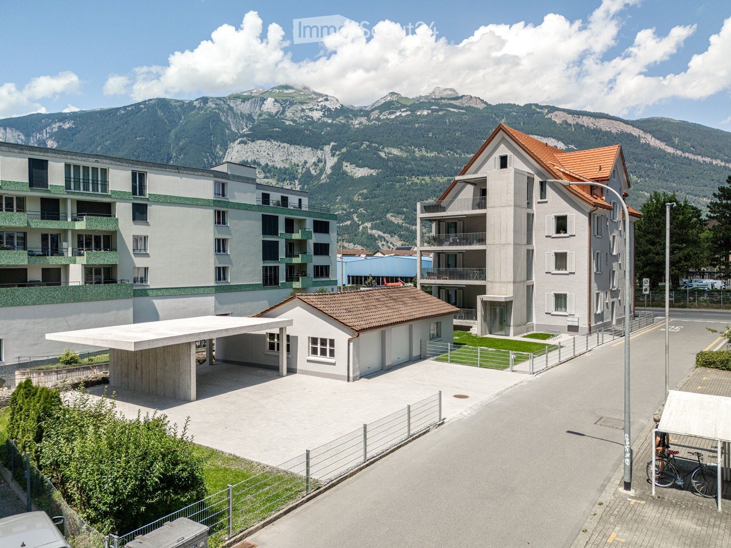 A multi-building residential complex with modern architecture, featuring a mix of apartment buildings and smaller houses, surrounded by a paved area, greenery, and a scenic mountain backdrop.
