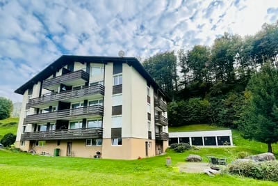 Multi-story residential building with balconies, surrounded by green lawn and trees.