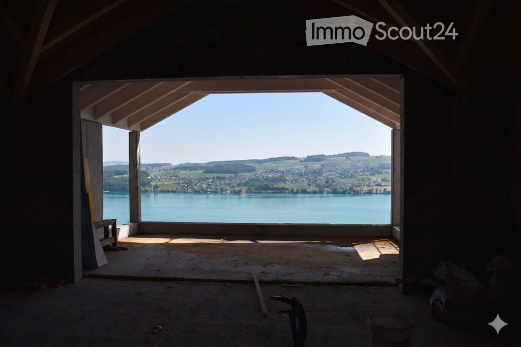Empty room with exposed wooden beams, large window offering a scenic view of a lake and surrounding landscape.