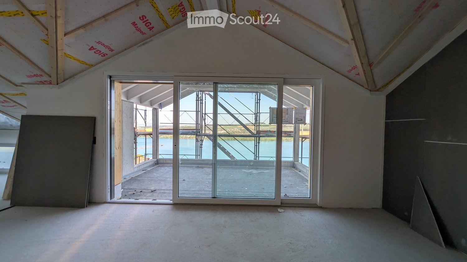 Empty room with exposed wooden beams, large glass sliding doors leading to an outdoor area with a view of water, and unfinished walls.
