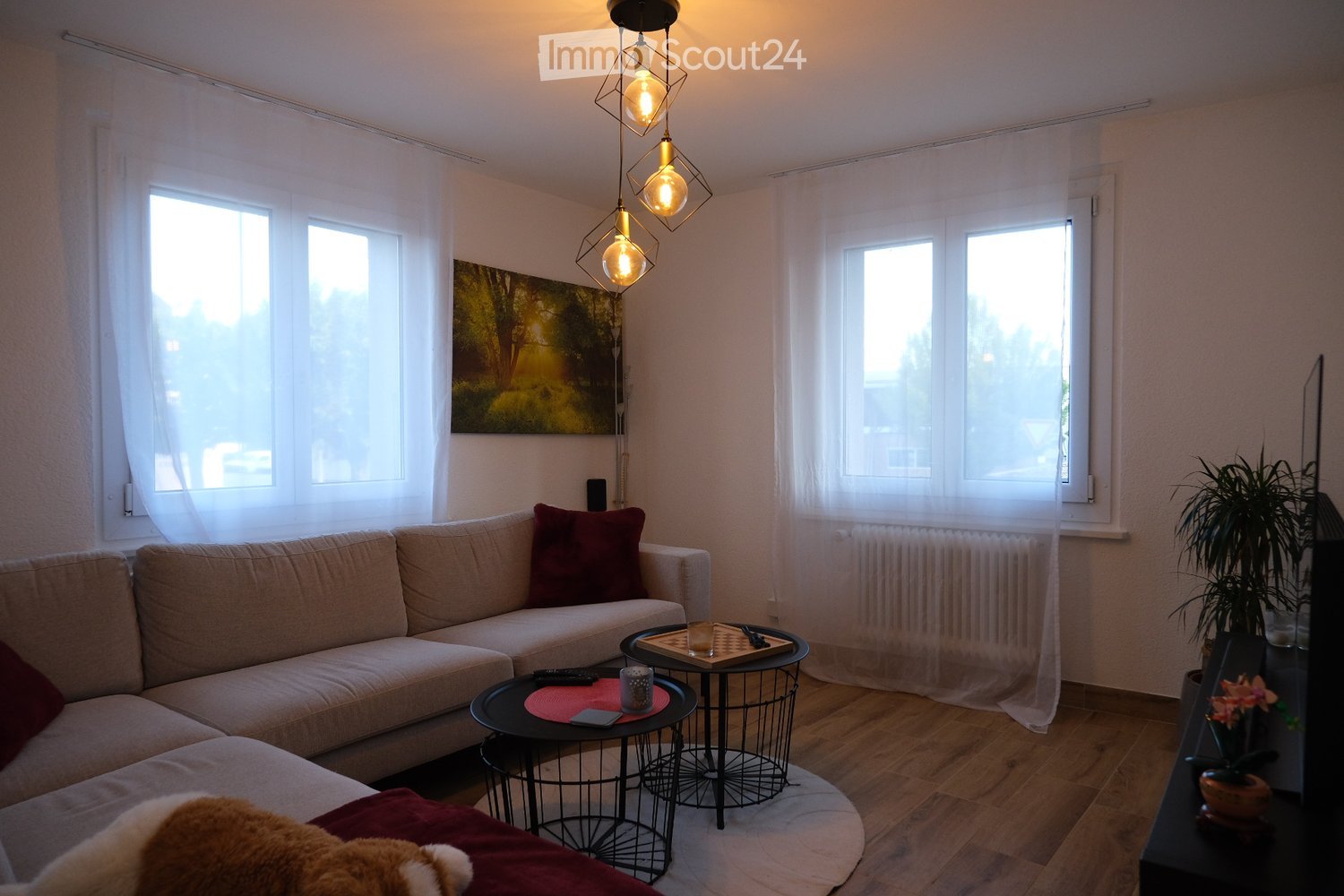 Modern living room with light-colored sofa, coffee table, side table, potted plant, and artwork on the wall. Large windows with white curtains allow natural light to fill the space.