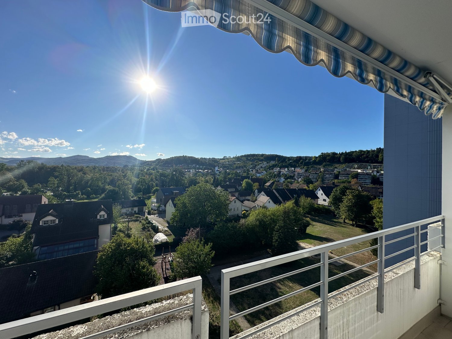 Balcony with a view of a town and surrounding landscape, featuring a striped awning and metal railing.