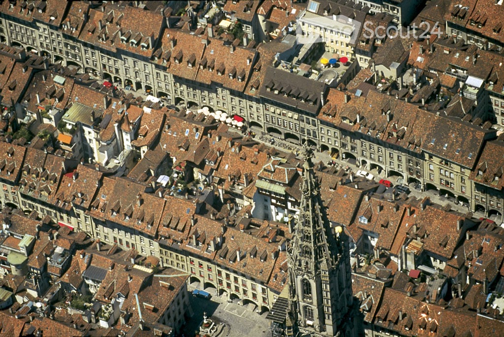 Aerial view of a historic European city with numerous buildings featuring red-tiled roofs, narrow streets, and a prominent church spire in the center.