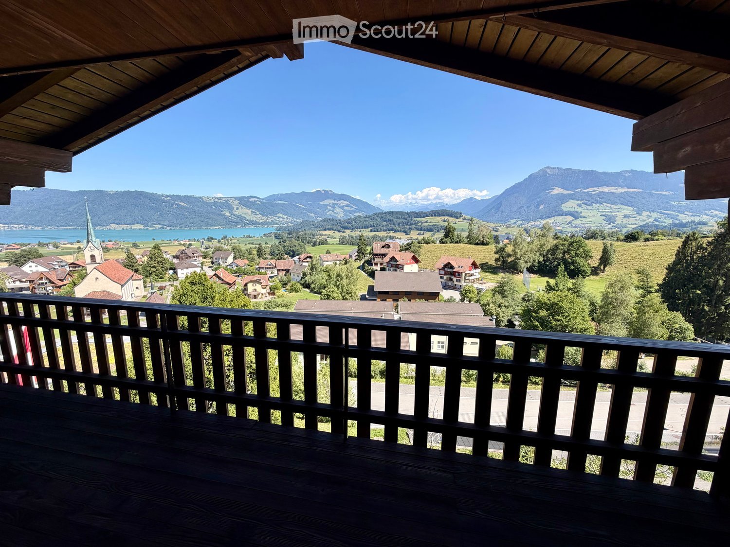 Balcony with wooden railing and a scenic view of a town, lake, and mountains.