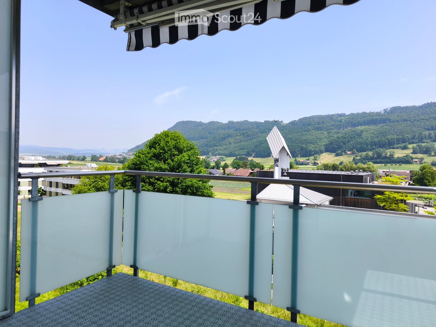 Balcony with glass railing, overlooking a scenic countryside with trees and hills under a clear blue sky.