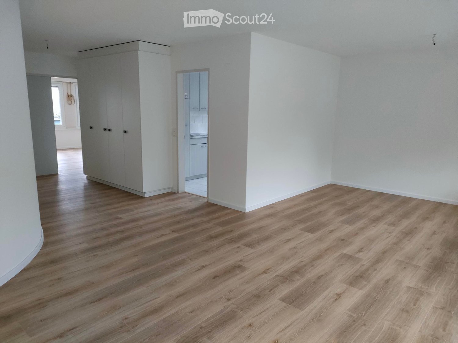 Empty room with light wooden flooring, white walls, and a built-in closet. The room is connected to a small kitchen area visible in the background.