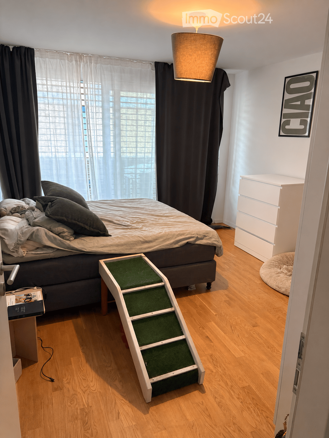 Cozy bedroom with a wooden floor, a large bed with pillows and blankets, a cat staircase, a white dresser, and a pet bed. Large window with sheer curtains and a decorative sign on the wall.