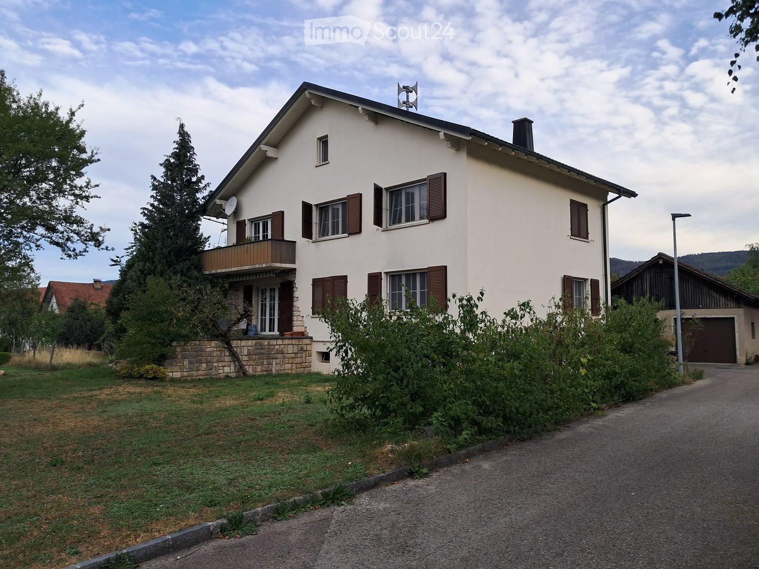 Two-story house with white exterior, brown shutters, and a balcony on the first floor. The house has a stone foundation and is surrounded by greenery and a driveway.