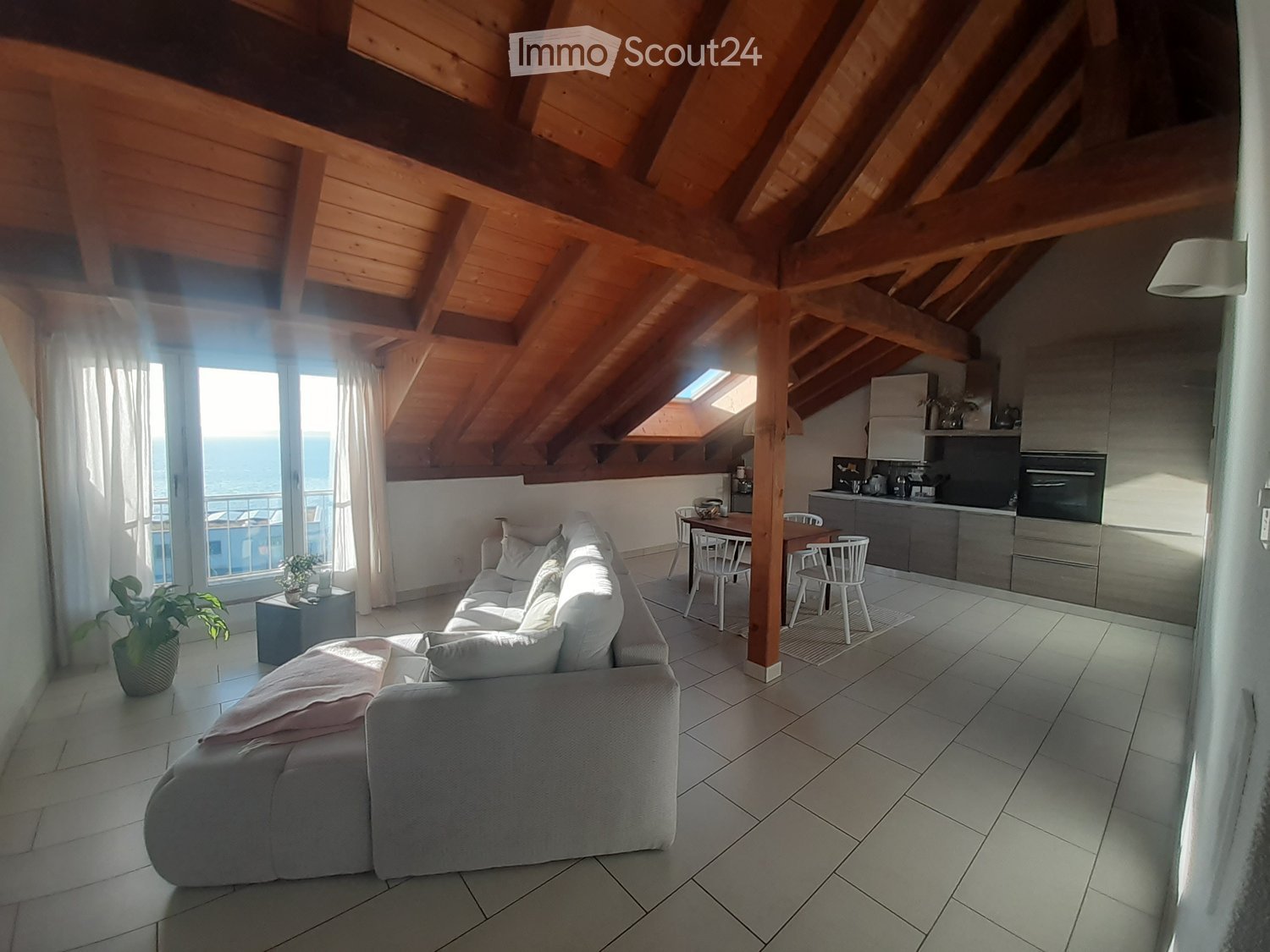 Bright, open living space with exposed wooden beams, a grey sectional sofa, a dining table with chairs, a modern kitchen area, and large windows offering a sea view.