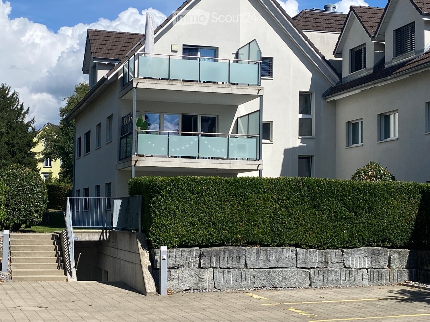 Modern two-story house with white exterior, multiple windows, balconies with glass railings, and a stone retaining wall in front.