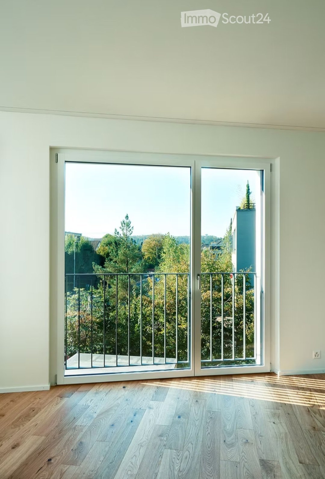 Empty room with light green walls, wooden flooring, and large sliding glass doors leading to a balcony with a metal railing and a view of trees and a building.