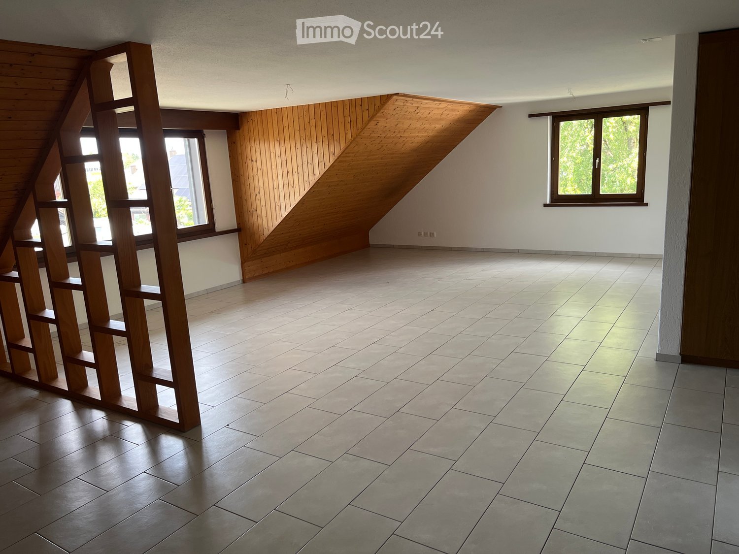 Empty room with wooden slatted shelving unit, tiled floor, and large windows with wooden frames.