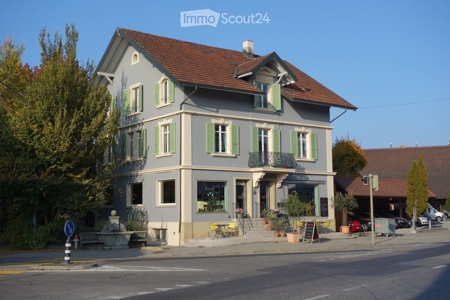 Two-story building with grey facade, green shutters, and a red-tiled roof. Features a small balcony, outdoor seating area, and a storefront with large windows.