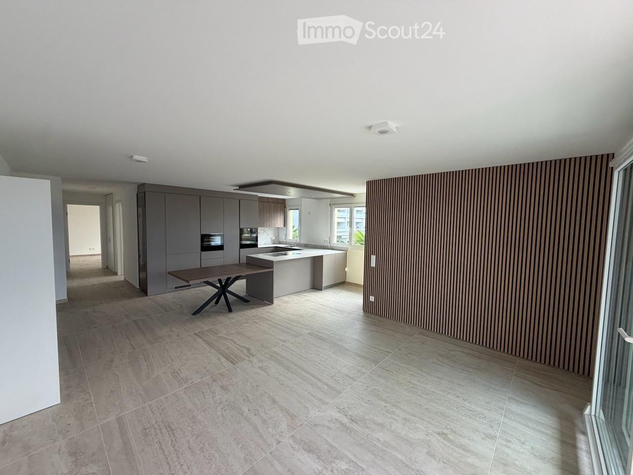 Modern kitchen and dining area with grey cabinets, a central island, a wooden dining table, and a sleek oven. The space features large windows and a contemporary design with a mix of wood and metal finishes.
