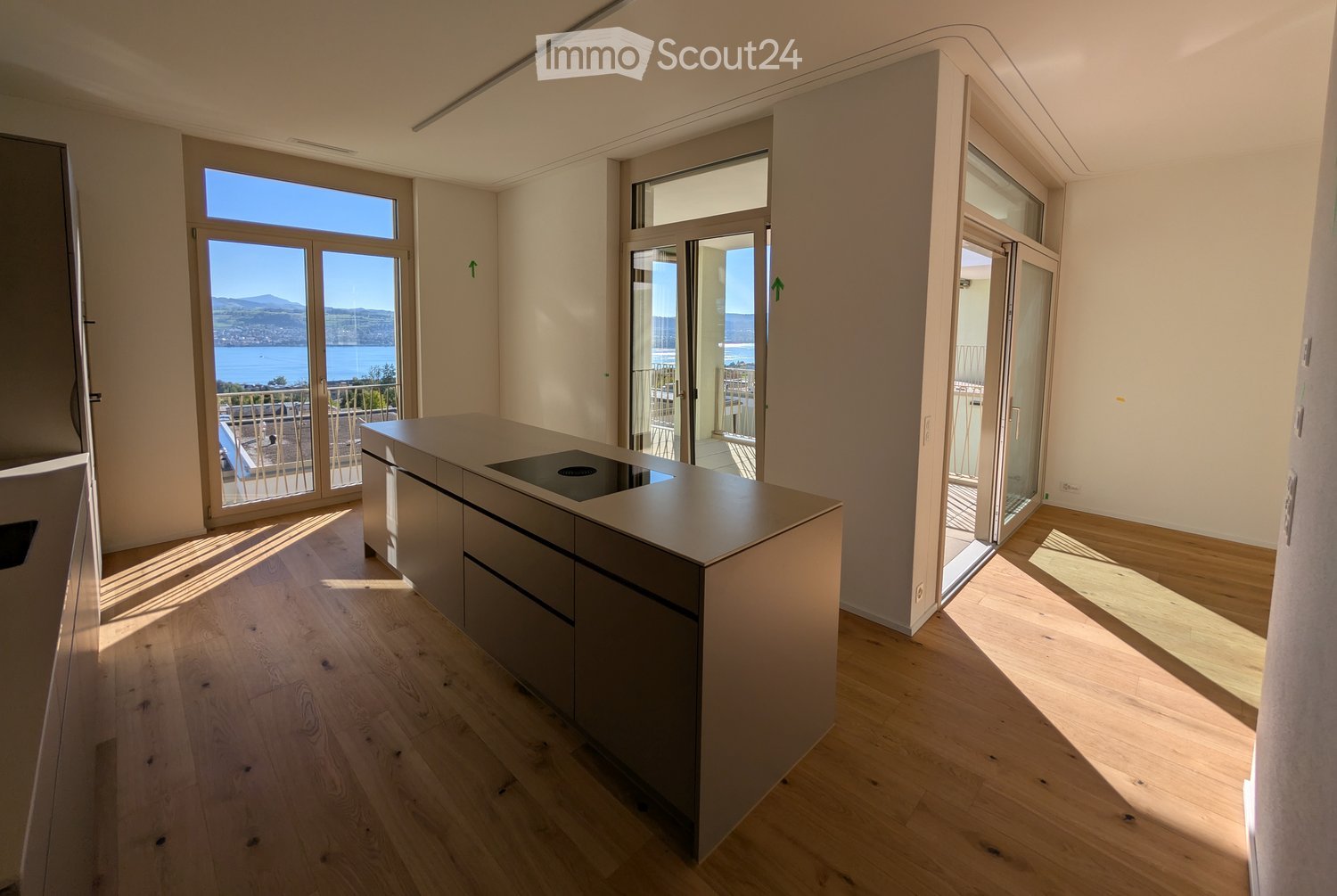 Modern kitchen with light wooden flooring, a central island with cabinets, and large windows offering a scenic view of the water and mountains.