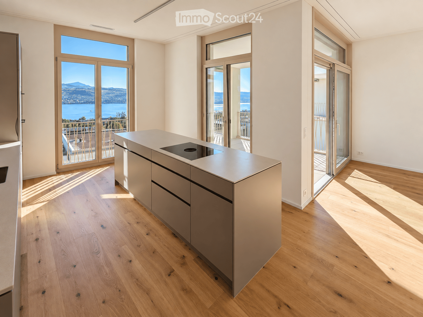 Modern kitchen with light wooden flooring, a central island with an induction cooktop, and large glass doors opening to a balcony with a scenic lake view.