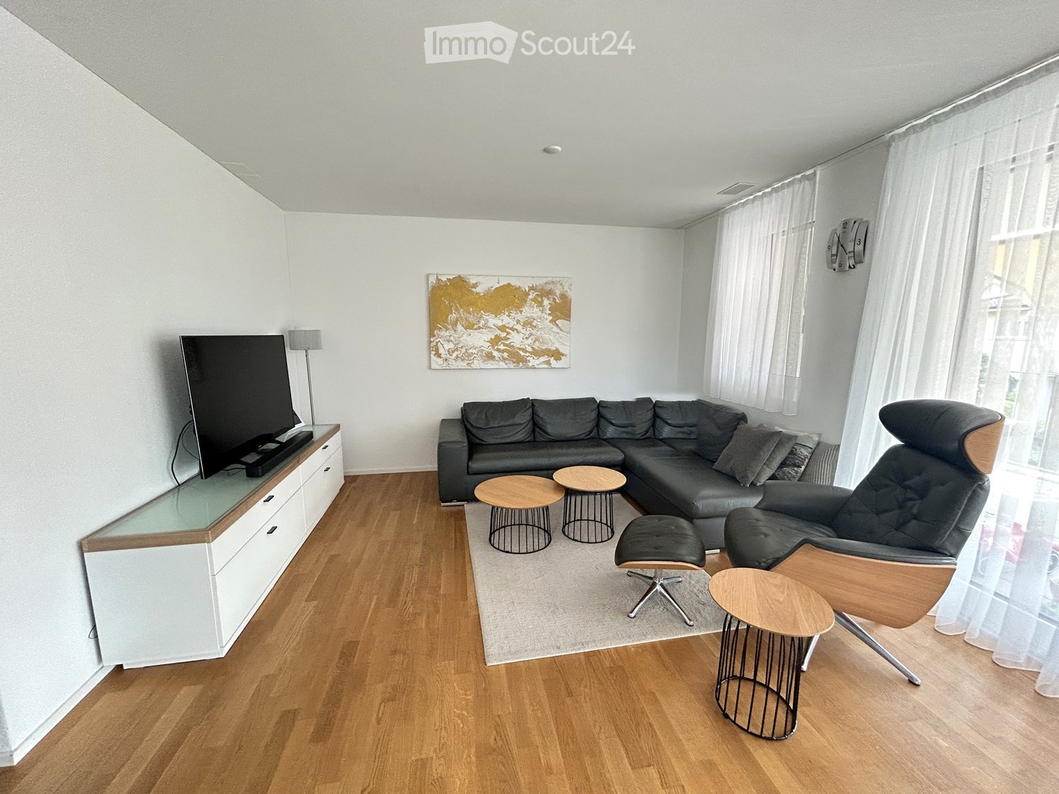 Modern living room with hardwood flooring, large sectional sofa, leather armchair, small round tables, TV on a white cabinet, and a decorative wall art piece.