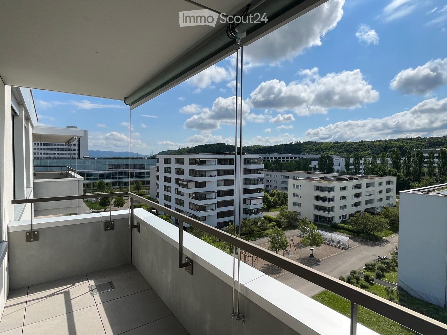 Balcony with modern metal railing, overlooking a campus-like setting with multiple white residential buildings, greenery, and a clear blue sky.