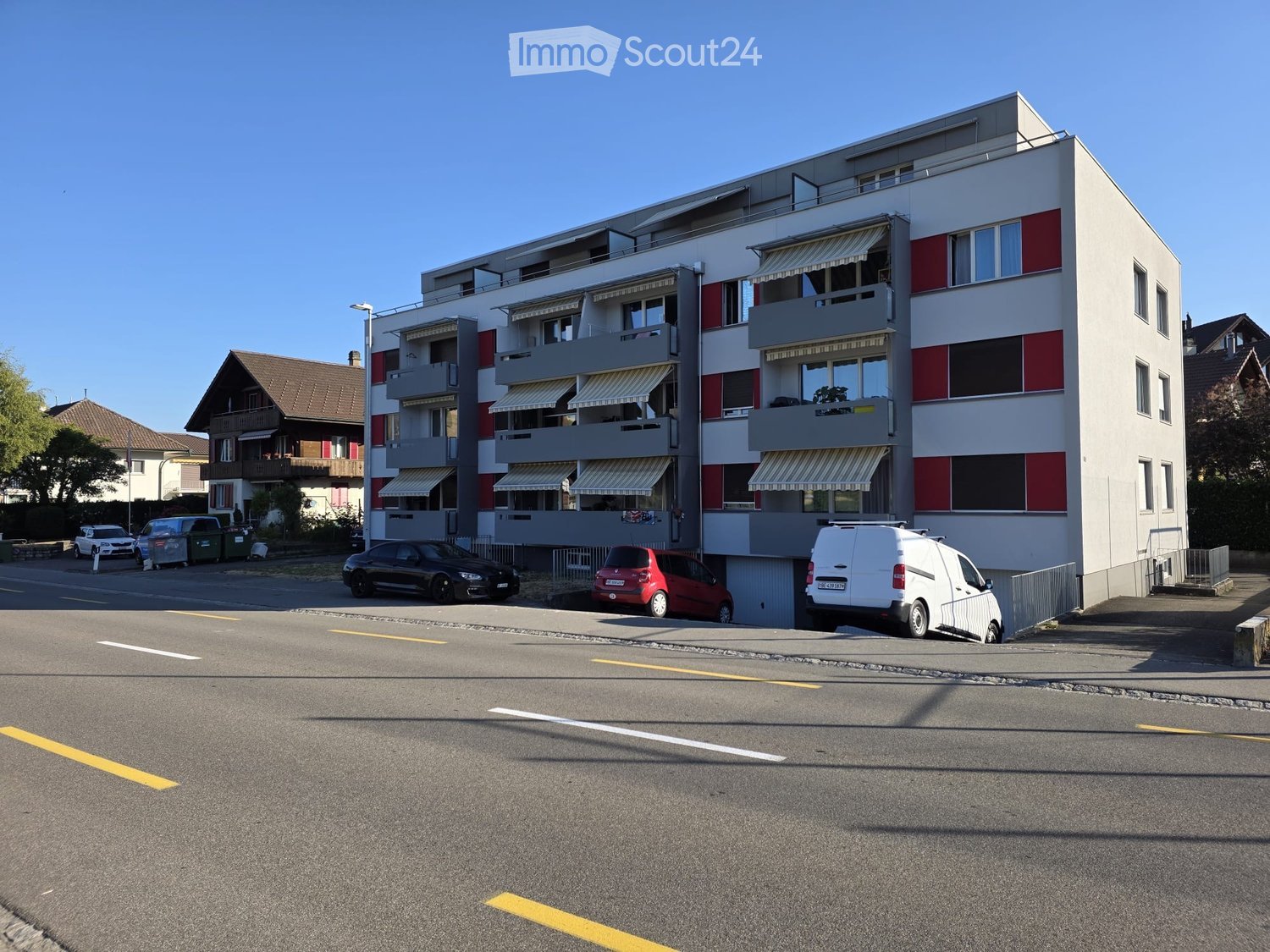 Modern multi-story apartment building with balconies, striped red and white facade, and several parked cars in front.