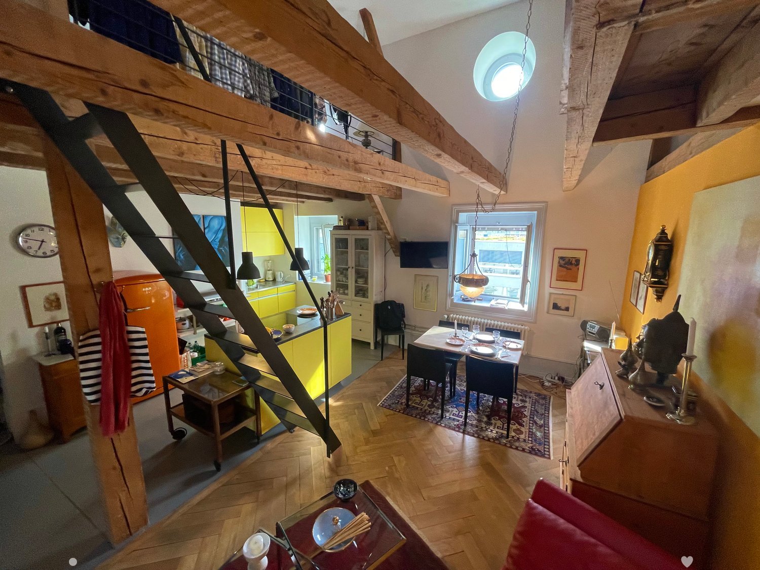 Open-plan living and dining area with wooden floors, exposed wooden beams, a staircase leading to a loft area, a red sofa, a glass coffee table, a dining table with chairs, a yellow kitchen area with cabinets and a refrigerator, and various decorative ite