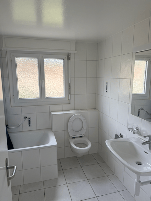 Modern bathroom with white tiled walls and floor, featuring a bathtub, toilet, sink, and mirror. The room has two windows with frosted glass panes, allowing natural light to enter.